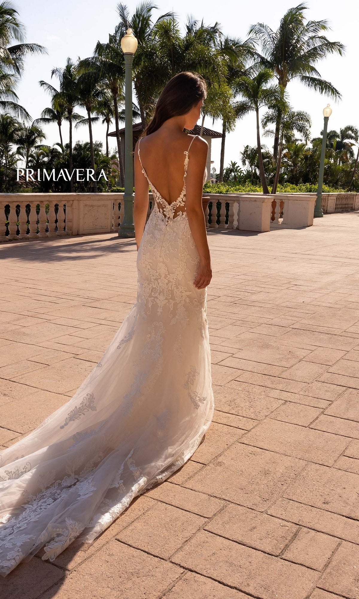 A woman wearing the Long Wedding Dress 11137 by Primavera—an ivory, fitted, lace-embroidered gown with a low back and long train—stands outdoors on a sunlit walkway lined with palm trees.